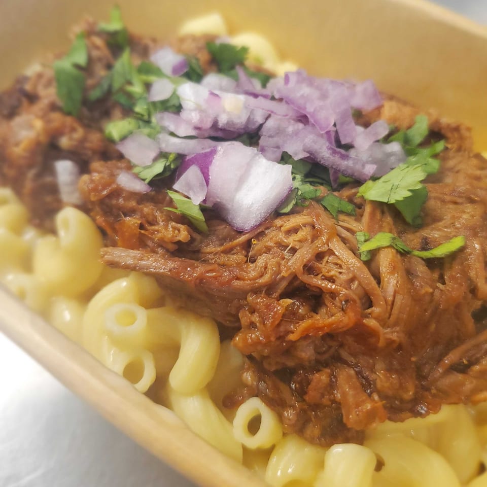 Pulled pork mac and cheese topped with red onions and cilantro in a brown takeout container.