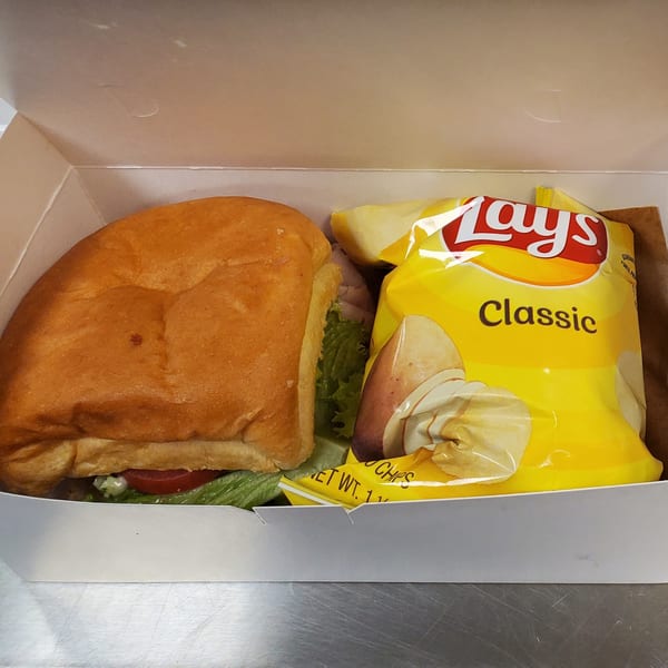 A Field House Deli sandwich with lettuce and tomato, next to a bag of Lay's Classic potato chips in a white box.