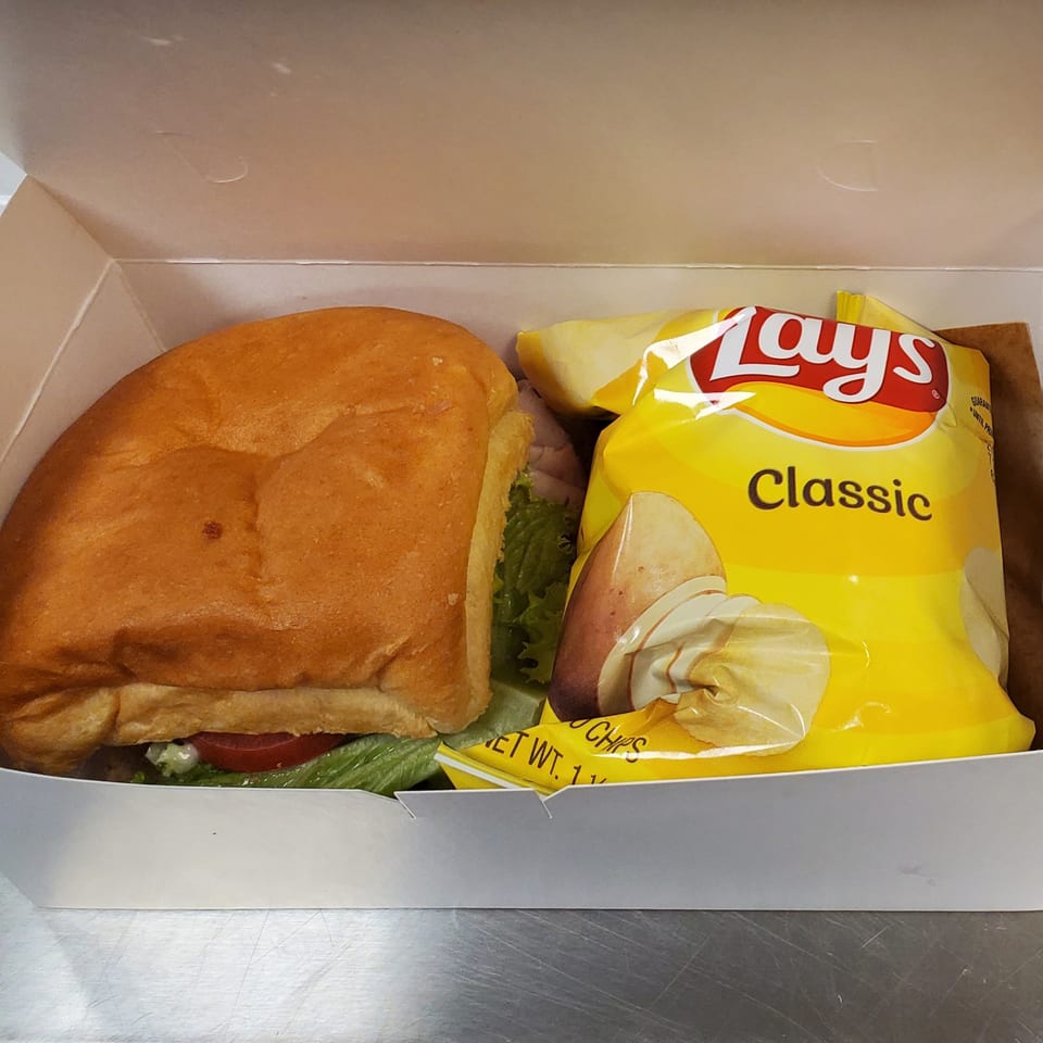 A Field House Deli sandwich with lettuce and tomato, next to a bag of Lay's Classic potato chips in a white box.