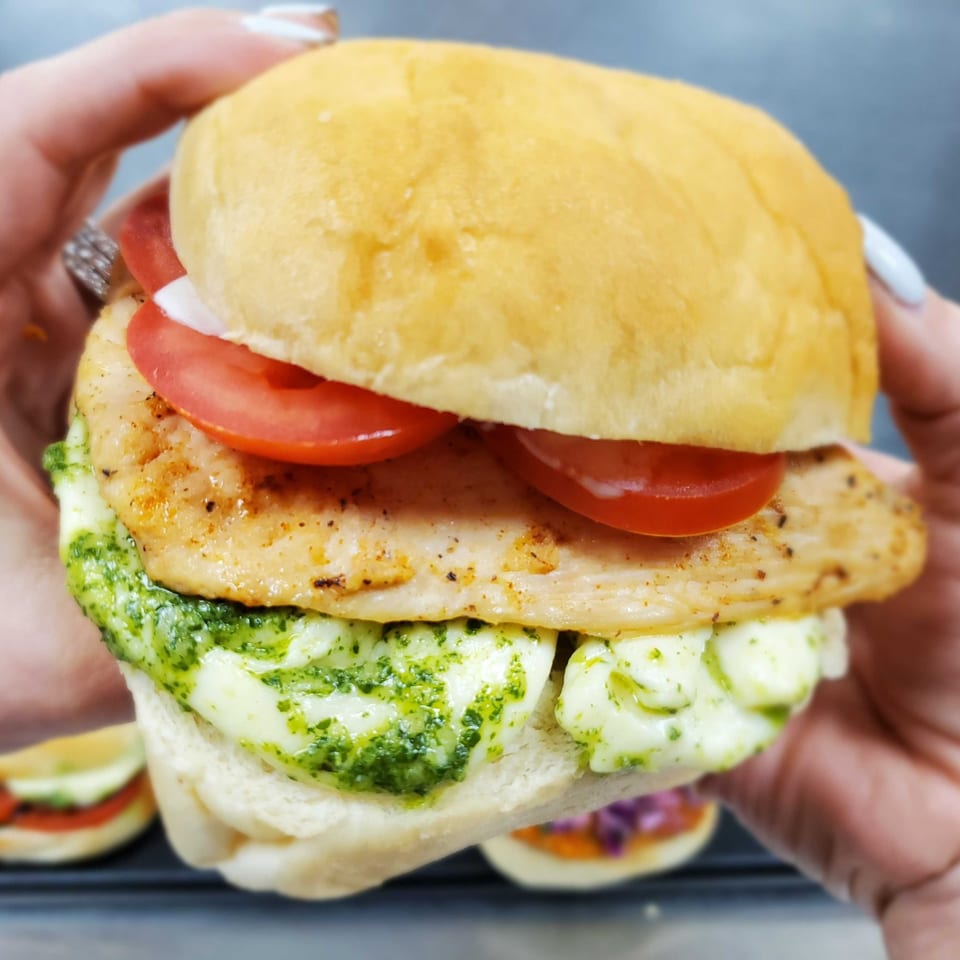 Hands holding a Field House Deli chicken sandwich with pesto, mozzarella, and tomatoes on a bun.
