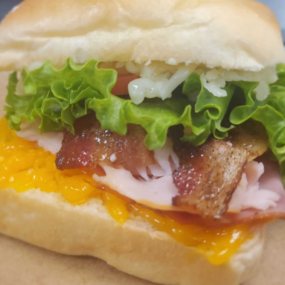 A Field House Deli sandwich with bacon, ham, melted cheese, lettuce, and mayonnaise on a bun.