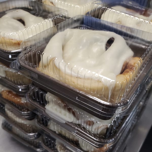 Several cinnamon rolls with white icing in clear plastic containers, stacked for Field House Deli.