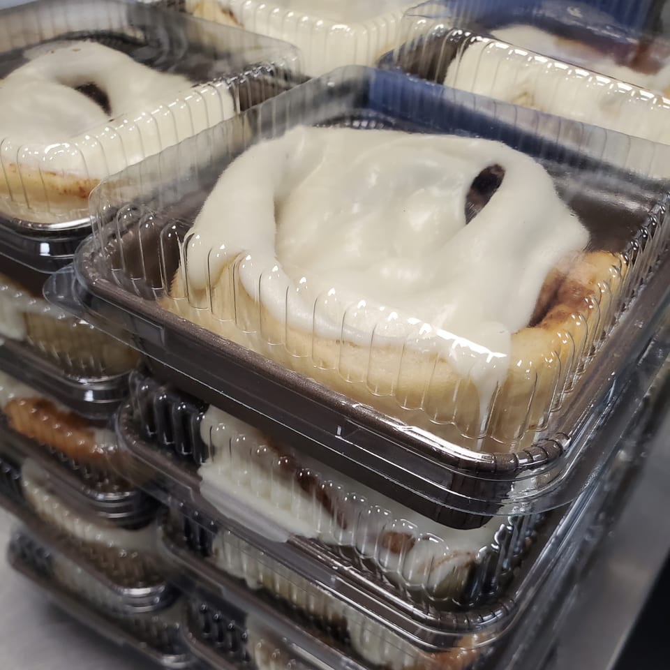 Several cinnamon rolls with white icing in clear plastic containers, stacked for Field House Deli.